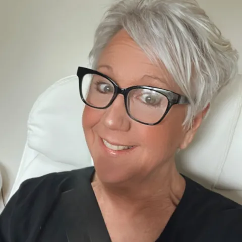 Mature Skin woman with short pixie like styled hair that is a silvery white. She’s wearing a black tee and has black framed glasses on. She has a big smile!