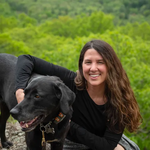 woman with long brown wavy hair in black shirt on green mountaintop with arm around black dog.