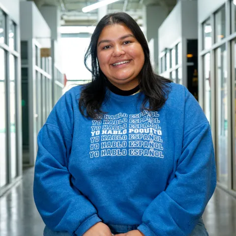 Helena smiling and sitting with her hands folded over each other over her lap, wearing a blue sweater that reads Yo Hablo Espanol multiple times in a row but in the middle in bold it says Yo Hablo Poquito 