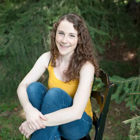 A smiling woman with long curly brown hair sits barefoot on a black wooden chair outdoors among evergreen branches. She wears a bright yellow tank top and blue jeans with her knees pulled up and her arms wrapped around her legs.
