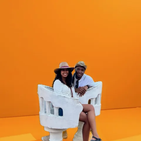 A man and a woman in white outfits sit on a white decorative chair against a bright orange background.