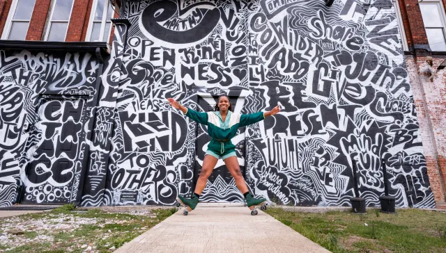 Person depicted on green roller skates pushing up on their heels with arms spread out against a black and white mural painted on a building 
