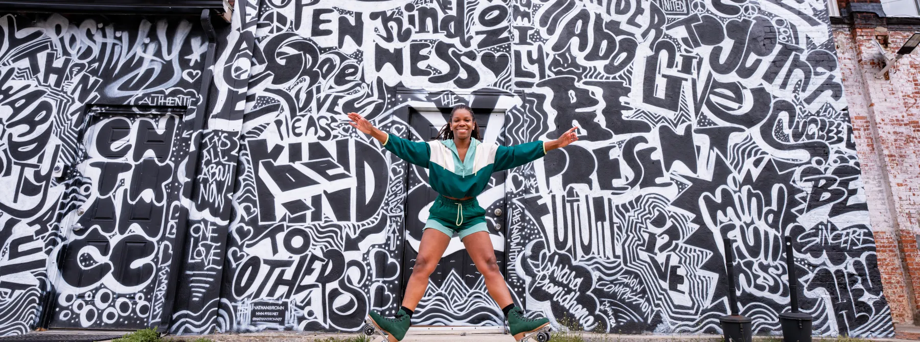 Person depicted on green roller skates pushing up on their heels with arms spread out against a black and white mural painted on a building 