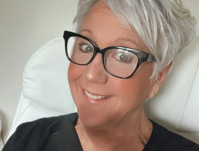 Mature Skin woman with short pixie like styled hair that is a silvery white. She’s wearing a black tee and has black framed glasses on. She has a big smile!