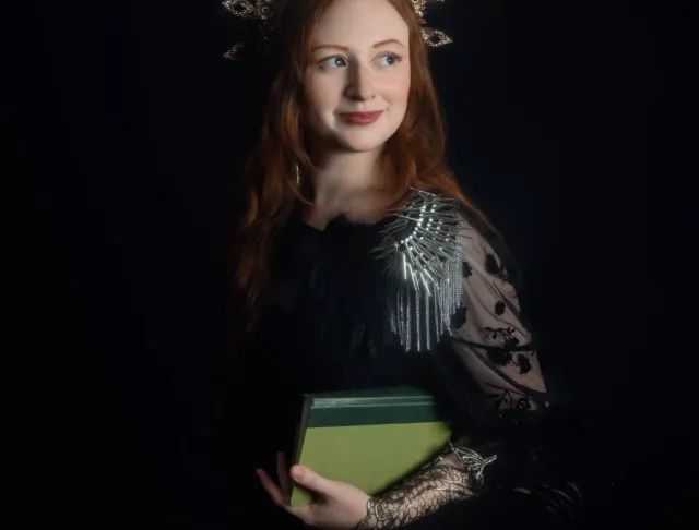 Kaitrin standing against a black background with a crown and a green book.