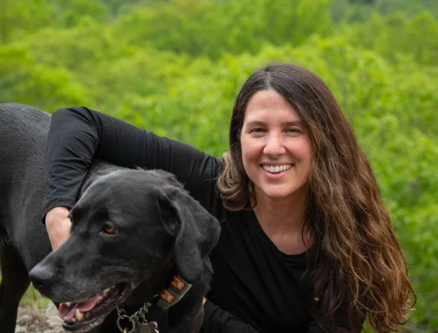 woman with long brown wavy hair in black shirt on green mountaintop with arm around black dog.