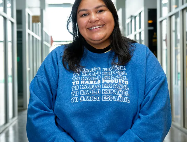 Helena smiling and sitting with her hands folded over each other over her lap, wearing a blue sweater that reads Yo Hablo Espanol multiple times in a row but in the middle in bold it says Yo Hablo Poquito 
