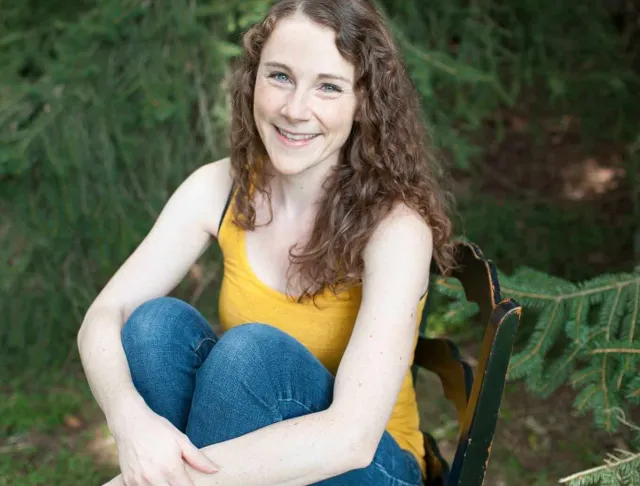 A smiling woman with long curly brown hair sits barefoot on a black wooden chair outdoors among evergreen branches. She wears a bright yellow tank top and blue jeans with her knees pulled up and her arms wrapped around her legs.