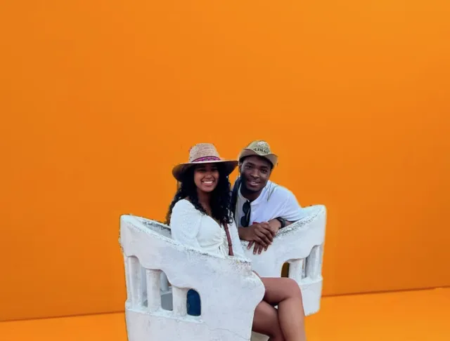 A man and a woman in white outfits sit on a white decorative chair against a bright orange background.
